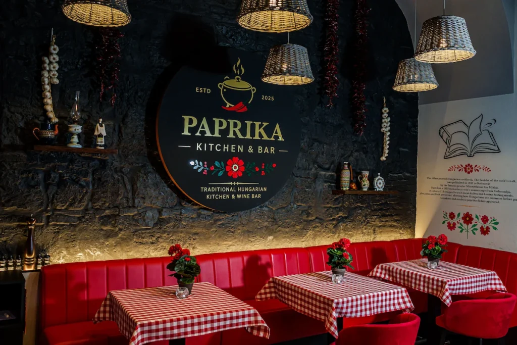 A Paprika Kitchen and Bar étterem hangulatos belső tere sötét kőfallal, kerek logóval, fonott lámpabúrákkal, fokhagymafüzérekkel és piros-fehér kockás asztalokkal.
